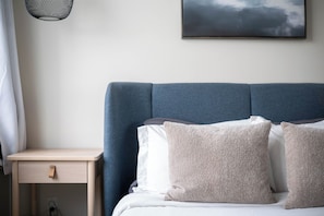 Een keurig opgemaakt bed met een blauw hoofdbord, een nachtkastje met lade en een stopcontact aan de muur.