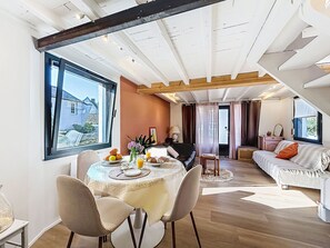 A dining area with a round table, chairs, and a view of a house through a large window. - Maison du Port by Interhome (Le Bono)