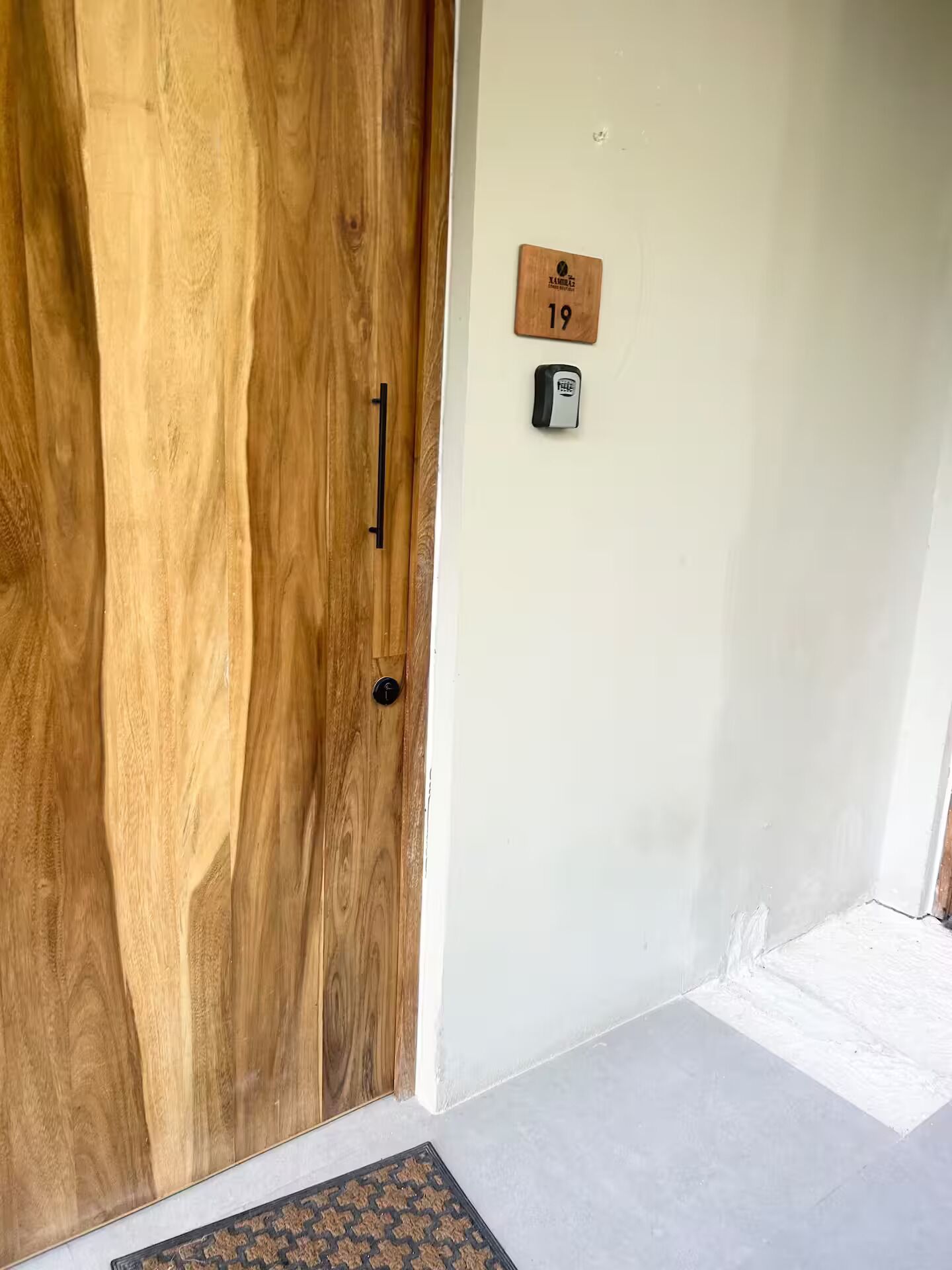A wooden door with a black handle, a small wooden plaque with the number '19', and a doorbell on the wall.