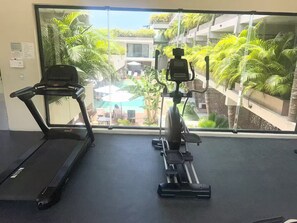 Ein Fitnessraum mit Laufband und Crosstrainer, große Fenster mit Blick auf einen Pool und Grünflächen.