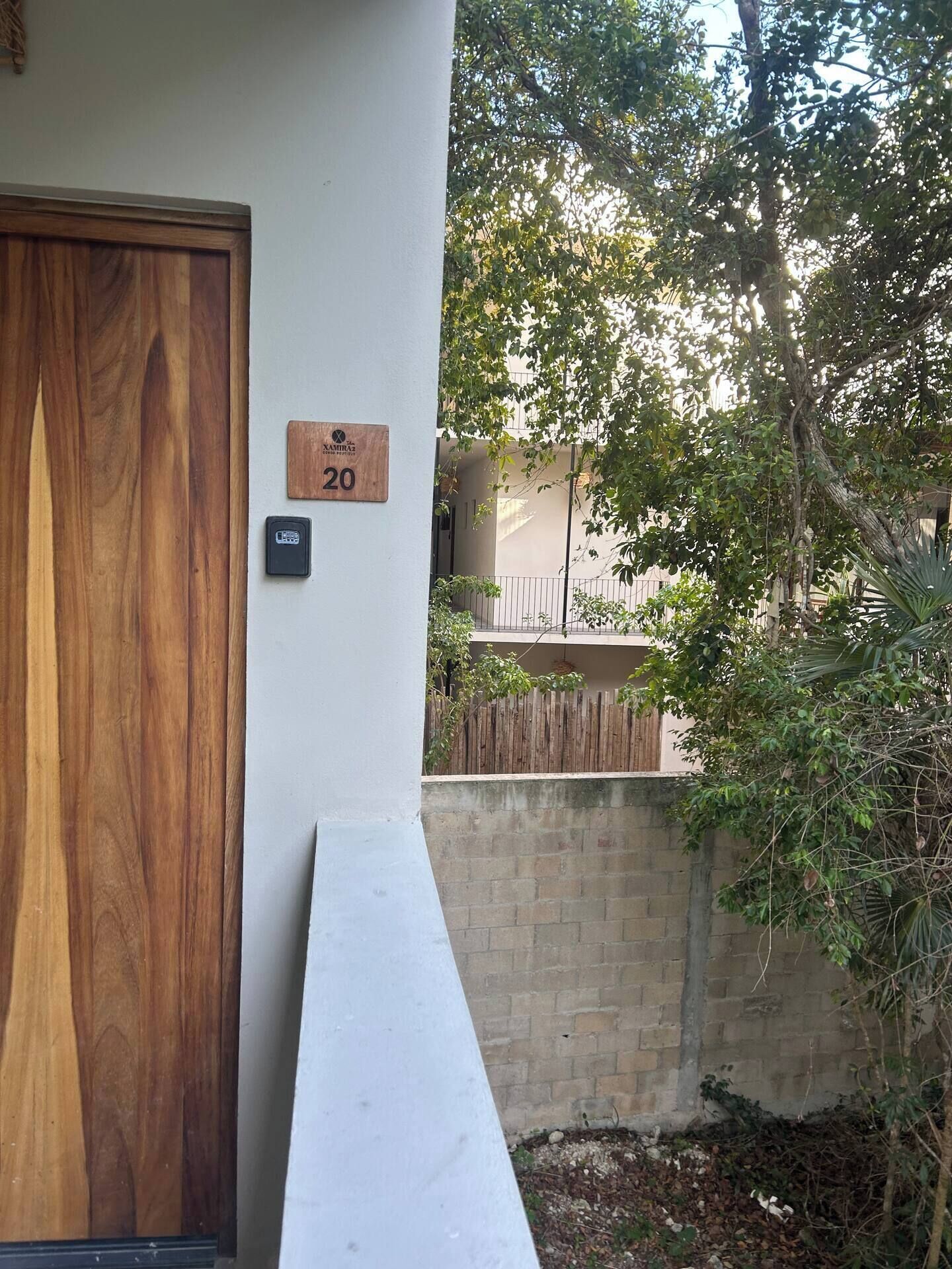 A wooden door with a number plaque, a keypad, and a white ramp leading to a gated area with trees and a building in the background.