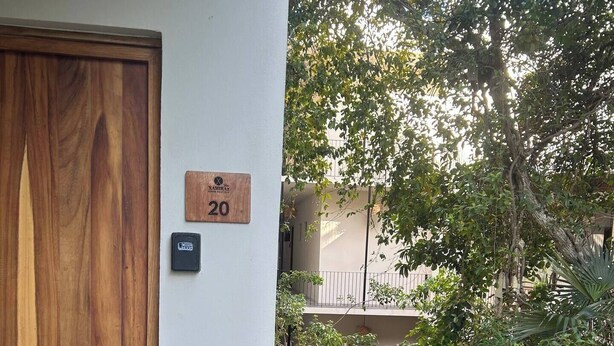 A wooden door with a number plaque, a keypad, and a white ramp leading to a gated area with trees and a building in the background. - ModernBlair Vacation Rentals (Tulum)