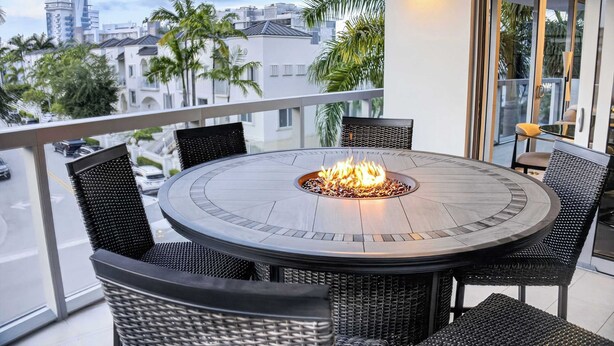A round outdoor dining area with a fire pit, surrounded by wicker chairs, on a balcony with a view of a cityscape. - Chic Bay Harbor 3BR • Rooftop Pool • Free Parking (Bay Harbor Islands)
