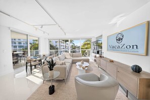 A modern living room with a sofa, coffee table, and a dining area with chairs and a glass table. There is a large window providing a view of buildings and palm trees. A framed sign on the wall reads 'VacaZon The Ultimate Vacation Location.' - Chic Bay Harbor 3BR • Rooftop Pool • Free Parking (Bay Harbor Islands)