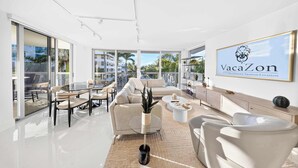 A modern living room with a glass dining table, comfortable seating, a coffee table, and a large window offering a view of palm trees and buildings. - Chic Bay Harbor 3BR • Rooftop Pool • Free Parking (Bay Harbor Islands)