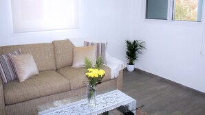 Ein Wohnzimmer mit einer Couch, einem Couchtisch mit Blumenvase, einer Topfpflanze und einem Fenster mit Jalousien.