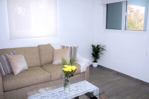 Un salón con un sofá, una mesa de centro con un jarrón de flores, una planta en maceta y una ventana con persianas.