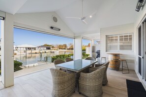 Uma área de refeições ao ar livre moderna, com mesa de vidro e cadeiras de vime, com vista para um corpo d'água e uma área residencial.