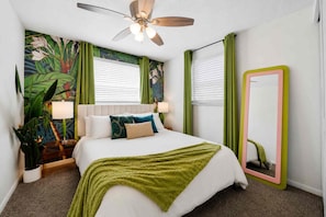 Une chambre à coucher comprenant un lit, un ventilateur de plafond, une table de chevet, un miroir et un mur orné d’une fresque aux motifs tropicaux.