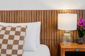 Uma cama bem arrumada com um travesseiro de padrão xadrez, uma luminária de cabeceira com cúpula de vidro transparente e um vaso com uma orquídea roxa.