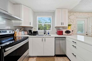 Uma cozinha moderna com armários brancos, fogão de aço inoxidável, lava-louças e uma janela com persianas.