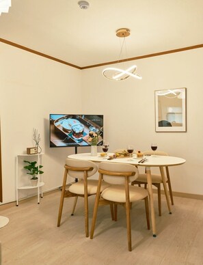 Ein Essbereich mit einem runden Tisch, vier Stühlen, einer kleinen Pflanze auf einem weißen Regal und einem an der Wand montierten Fernseher.