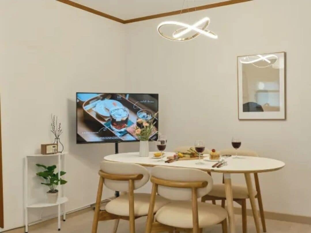 A dining area with a round table, four chairs, a television mounted on the wall, and a small plant on a shelf.