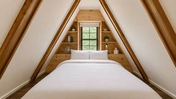 Ein Schlafzimmer mit schrägen Wänden, Holzbalken, einem Bett mit weißer Bettwäsche, einem Fenster und einer kleinen Ablage mit Gegenständen.
