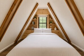 Ein Schlafzimmer mit schrägen Wänden, Holzbalken, einem Bett mit weißer Bettwäsche, einem Fenster und einer kleinen Ablage mit Gegenständen.