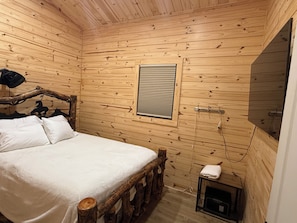 Ein Zimmer in einer Holzhütte mit einem Bett, einem Fenster mit Jalousien, einem kleinen Tisch und einem Heizgerät.
