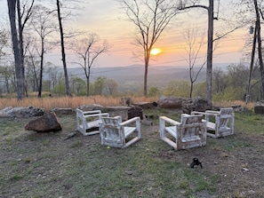 A sunset view with four rustic wooden chairs arranged on a grassy area, surrounded by trees and rocks.