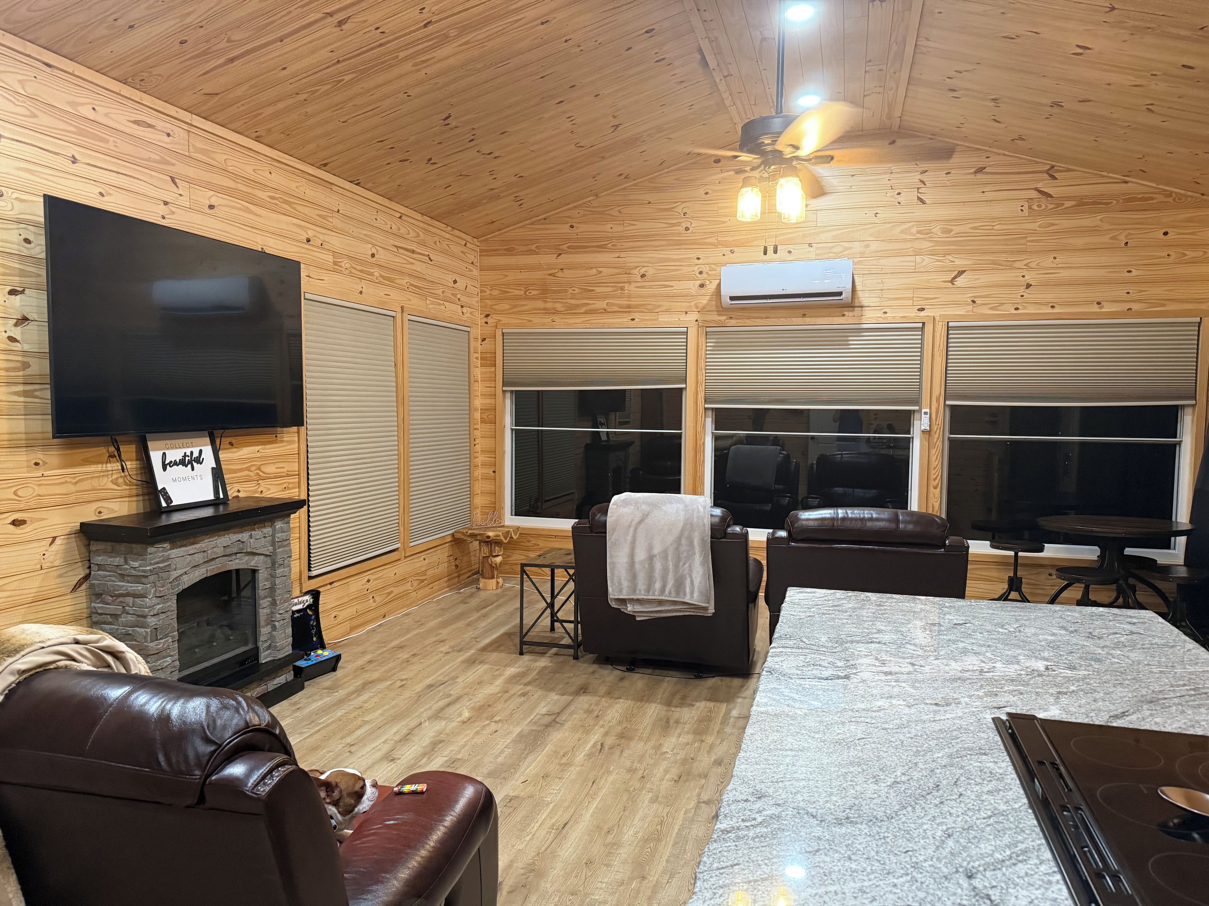 A cozy cabin interior with a fireplace, leather seating, a flat-screen TV, and a ceiling fan.