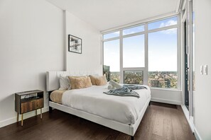 Quarto moderno com uma janela ampla, uma cama com roupa de cama branca, uma mesa de cabeceira e um quadro na parede.