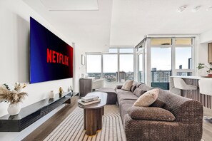 Uma sala de estar moderna com uma grande TV de tela plana exibindo 'NETFLIX' em letras vermelhas, um conjunto de sofás, uma mesa de centro e vista da cidade através de grandes janelas.