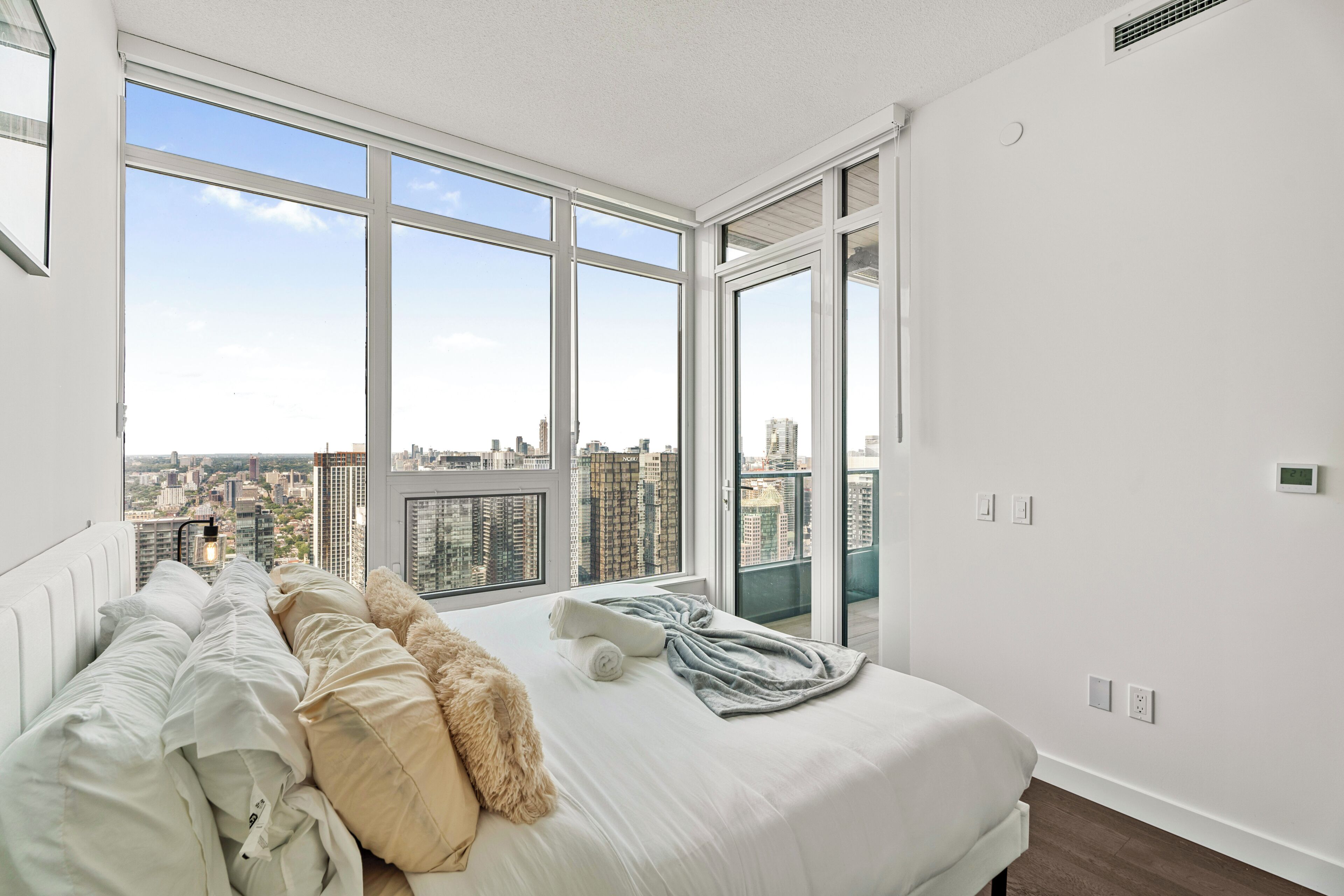 Ein Schlafzimmer mit einem großen Bett, Blick auf eine Stadtlandschaft durch raumhohe Fenster und einer weißen Wand mit Thermostat.