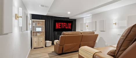 A modern living room with a flat-screen TV, leather recliner sofas, a wooden cabinet, and a wall-mounted shelf.