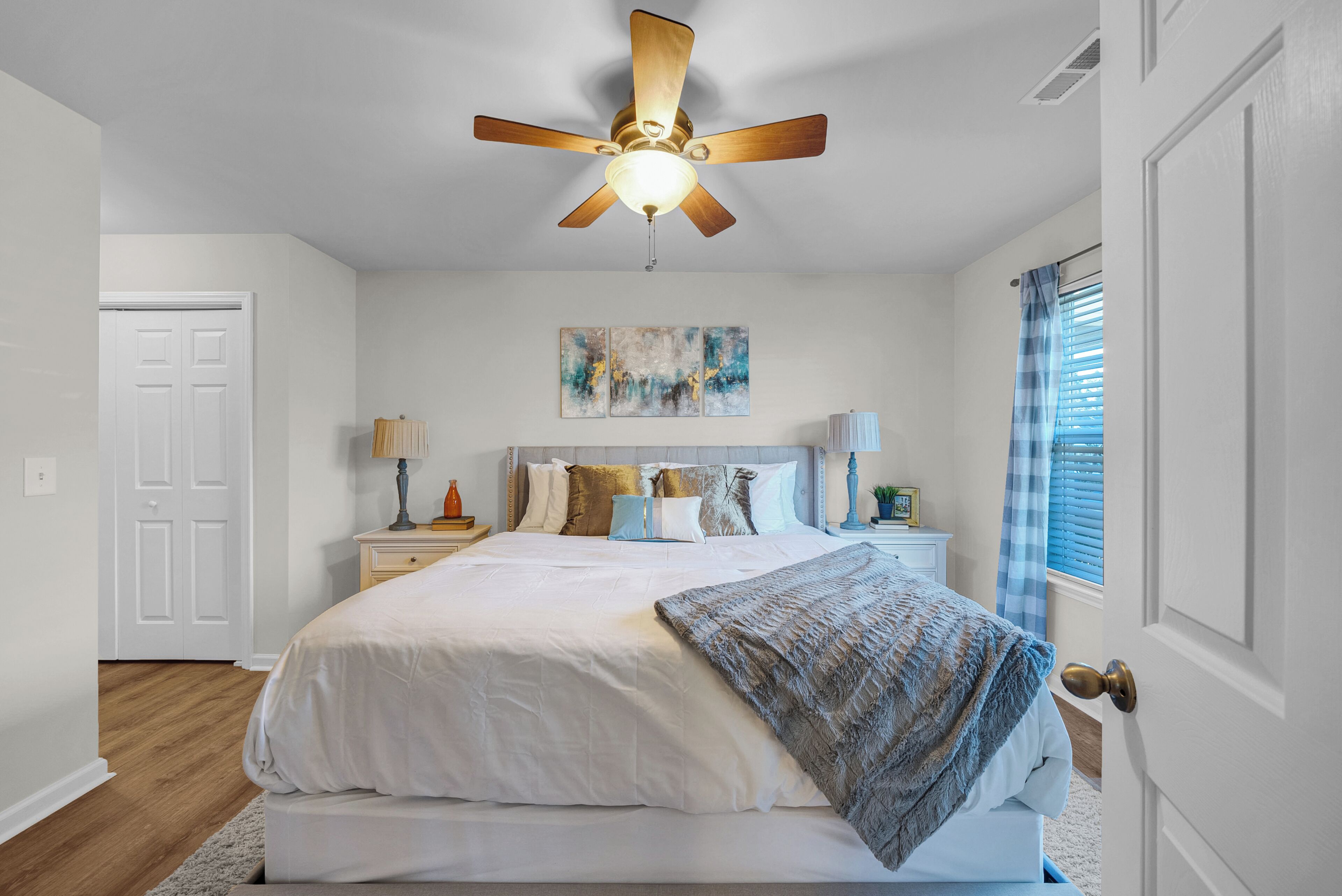 Ein Schlafzimmer mit Bett, Deckenventilator, Nachttisch, Lampe, Fenster mit Vorhängen und einer Tür.