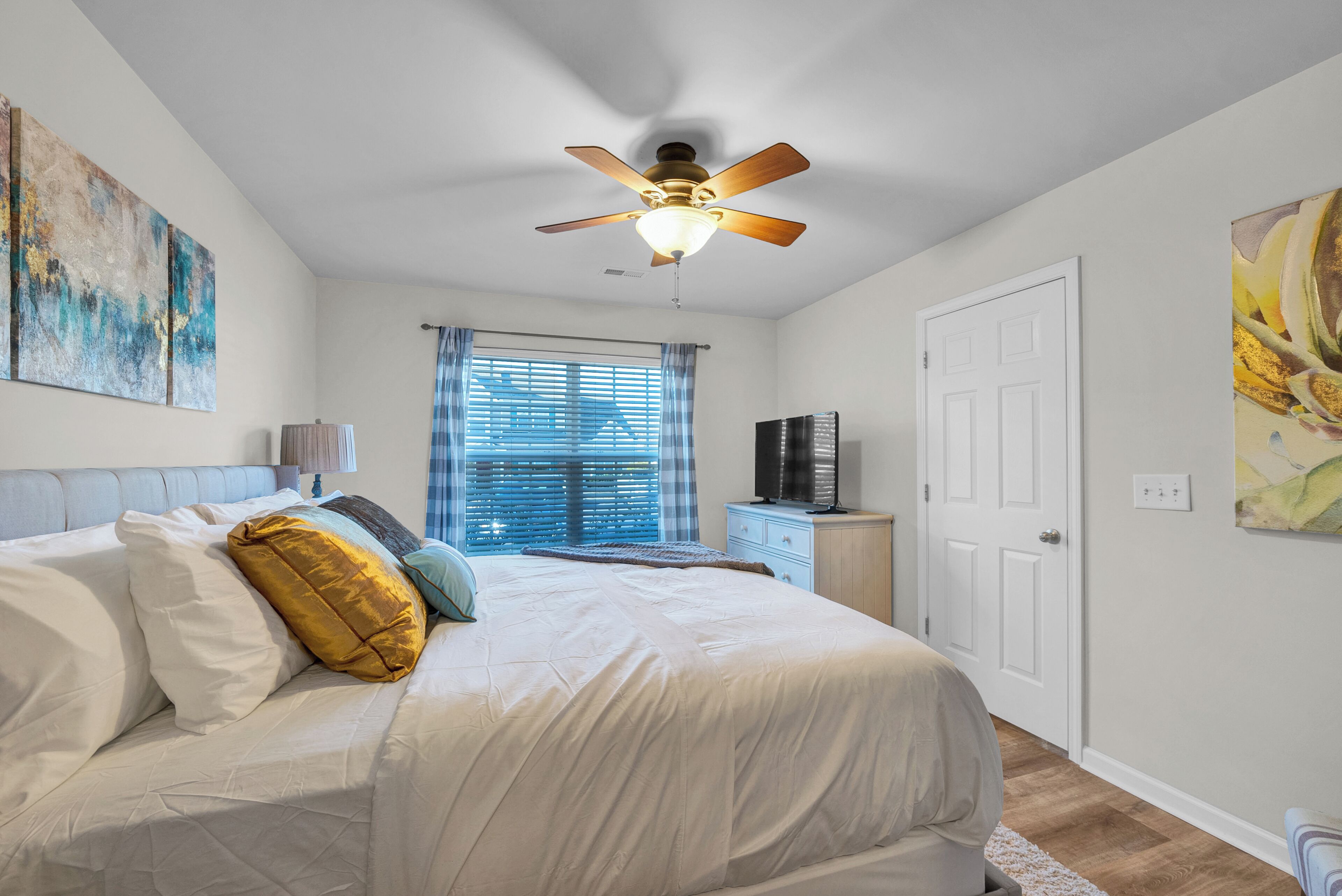 Une chambre à coucher avec un lit, un ventilateur de plafond, une fenêtre avec des stores, une porte et un tableau accroché au mur.