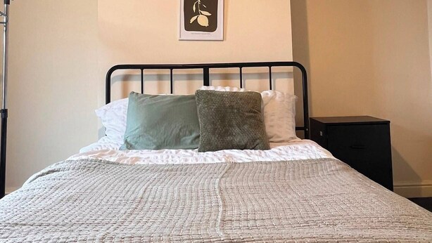 Un dormitorio con cama, almohadas, un cuadro en la pared y una mesita de noche.