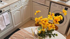 Una mesa redonda de comedor con mantel beige, dos platos con motivos florales, cubiertos dorados y un florero con flores amarillas.