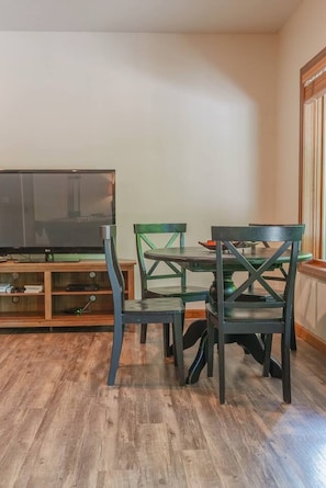 A dining area with a wooden table and chairs, a television on a stand, and a window with blinds. - Travis Lodge | Hot Tub, Game Room & Trails (Logan)