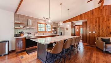 A modern kitchen with a central island, bar stools, wooden flooring, and pendant lighting.