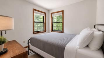 A bedroom with a bed, two windows, a bedside table with a lamp, and a small plant.