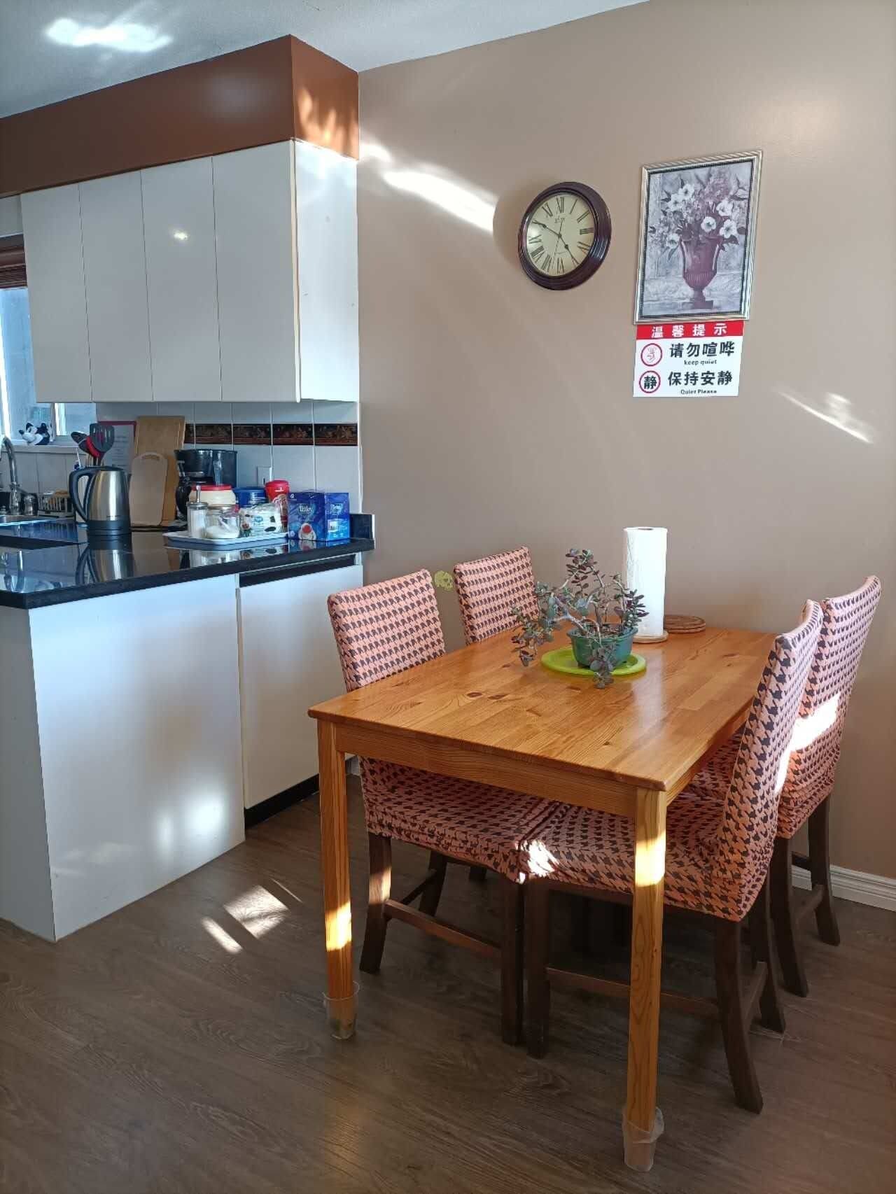A dining area with a wooden table and patterned chairs, a clock on the wall, and a kitchen in the background.