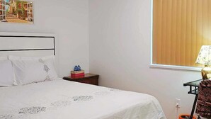 Een slaapkamer met een bed, een nachtkastje, een raam met jaloezieën, een lamp en een kleine tafel met een waterfles.