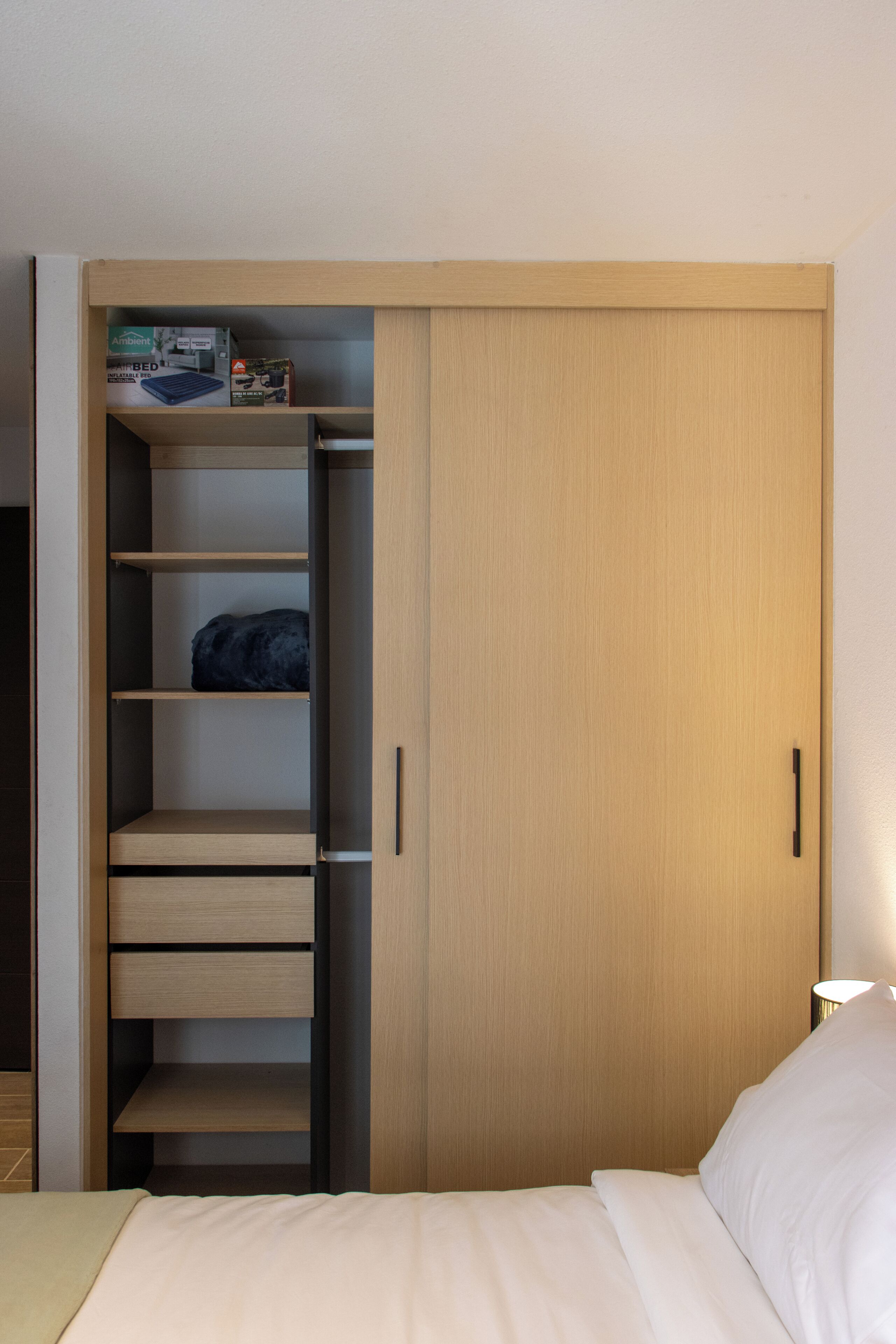 A bedroom with a wooden wardrobe, a bed with white bedding, and a nightstand with a lamp.
