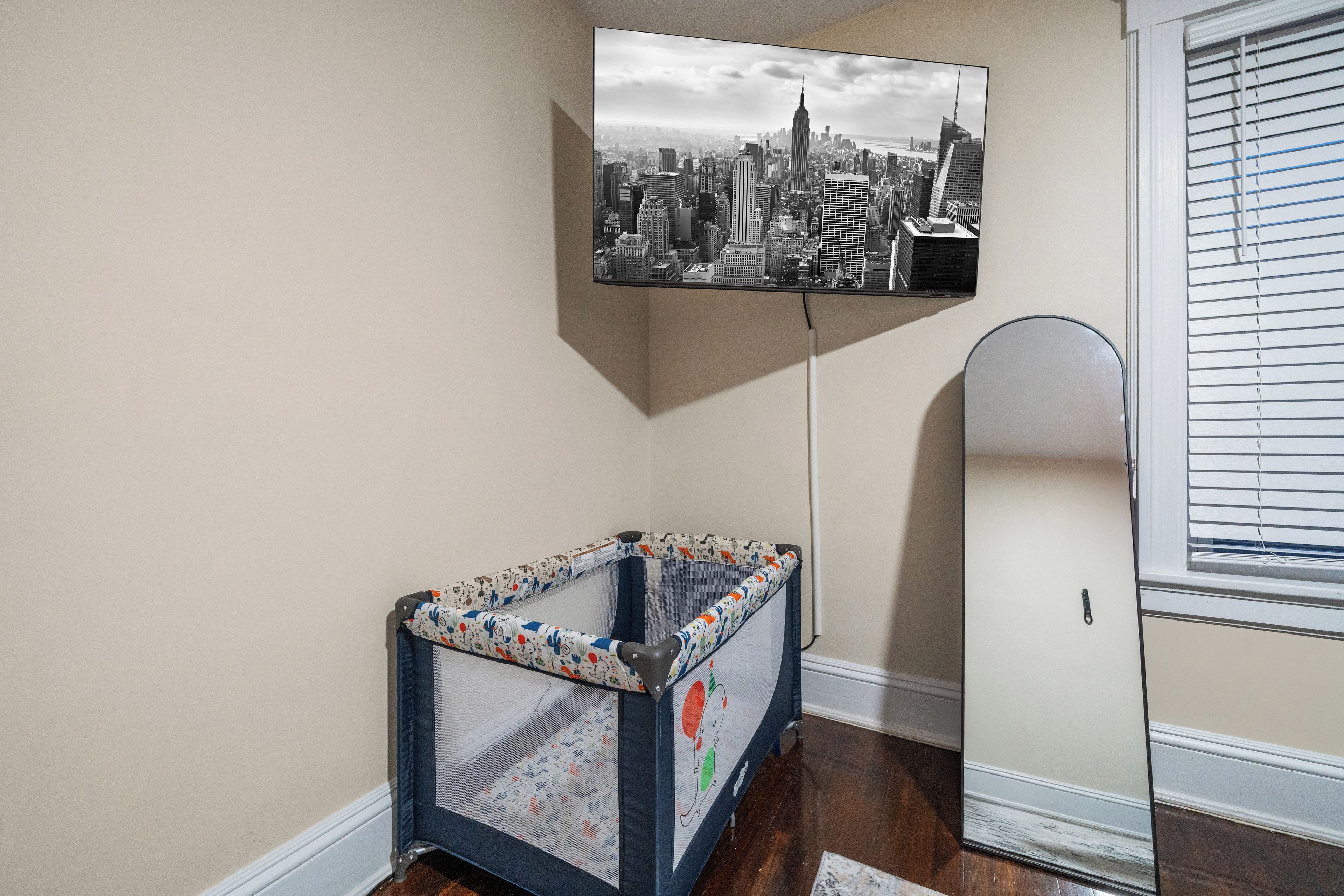 Habitación con televisión mostrando un paisaje urbano, un corralito infantil en una esquina con un diseño llamativo y una ventana con persianas.