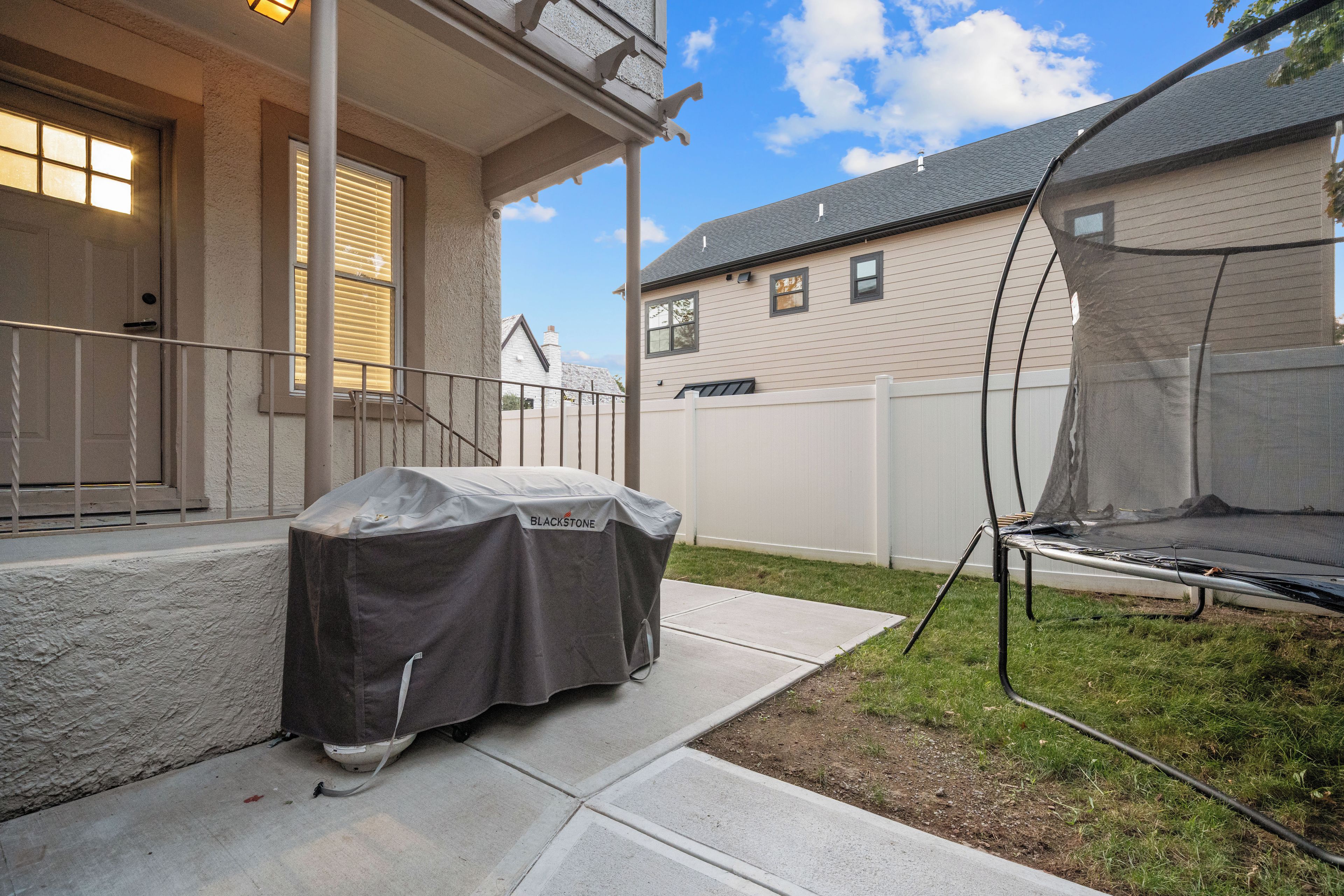 Una parrilla exterior techada, un trampolín y una casa con valla blanca al fondo.