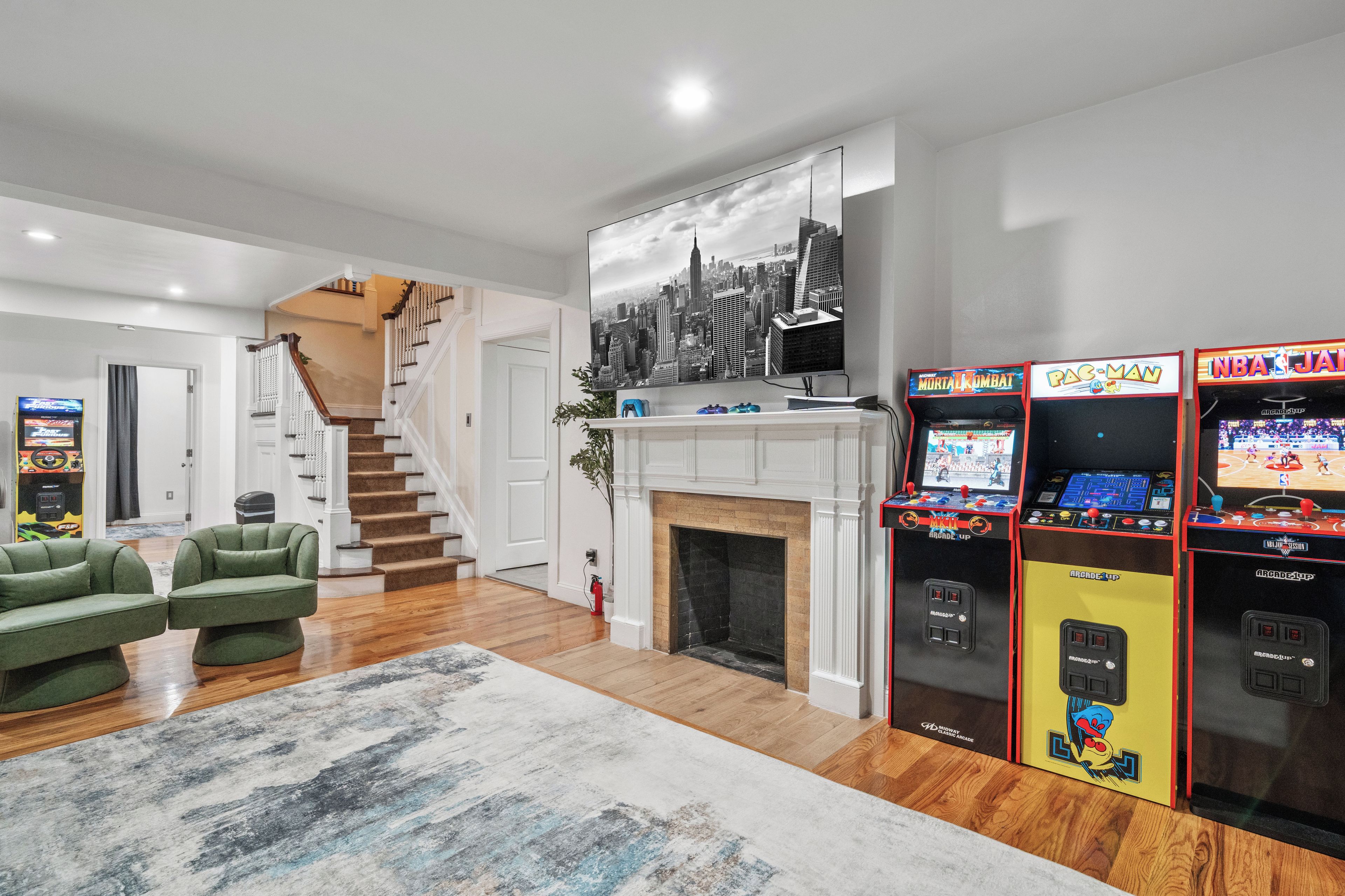 A living room with a fireplace, green sofas, a staircase, and arcade games.