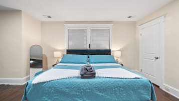 Una cama bien hecha con edredón azul y blanco, una lámpara de noche y un espejo a la izquierda.