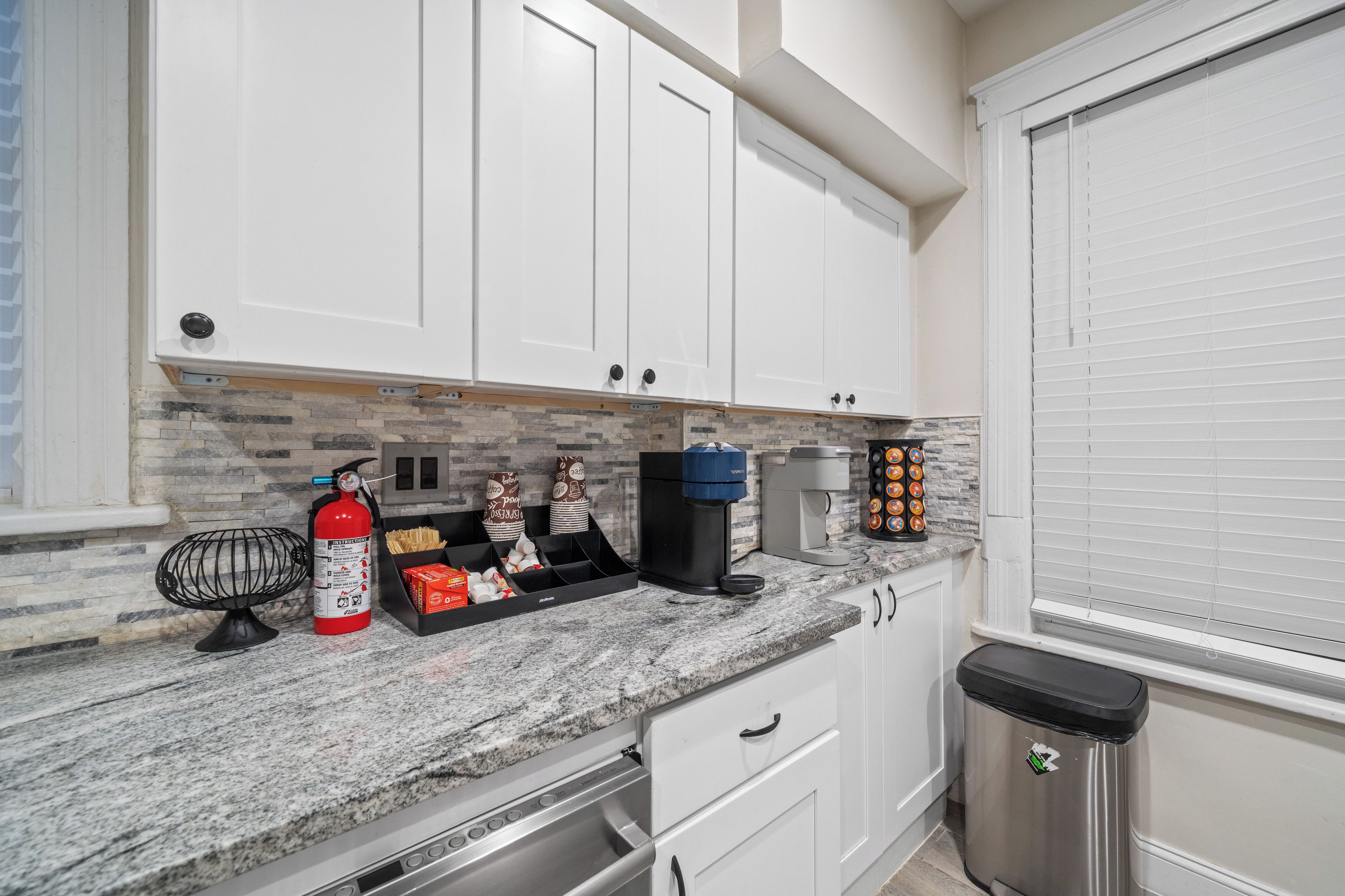 A kitchen with white cabinets, a granite countertop, a coffee maker, and a trash can.