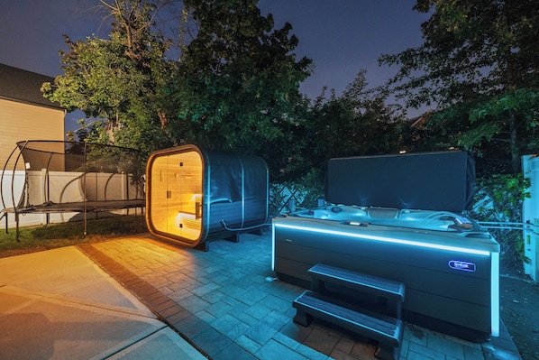 An outdoor sauna with a bench and a table, surrounded by trees and a house.