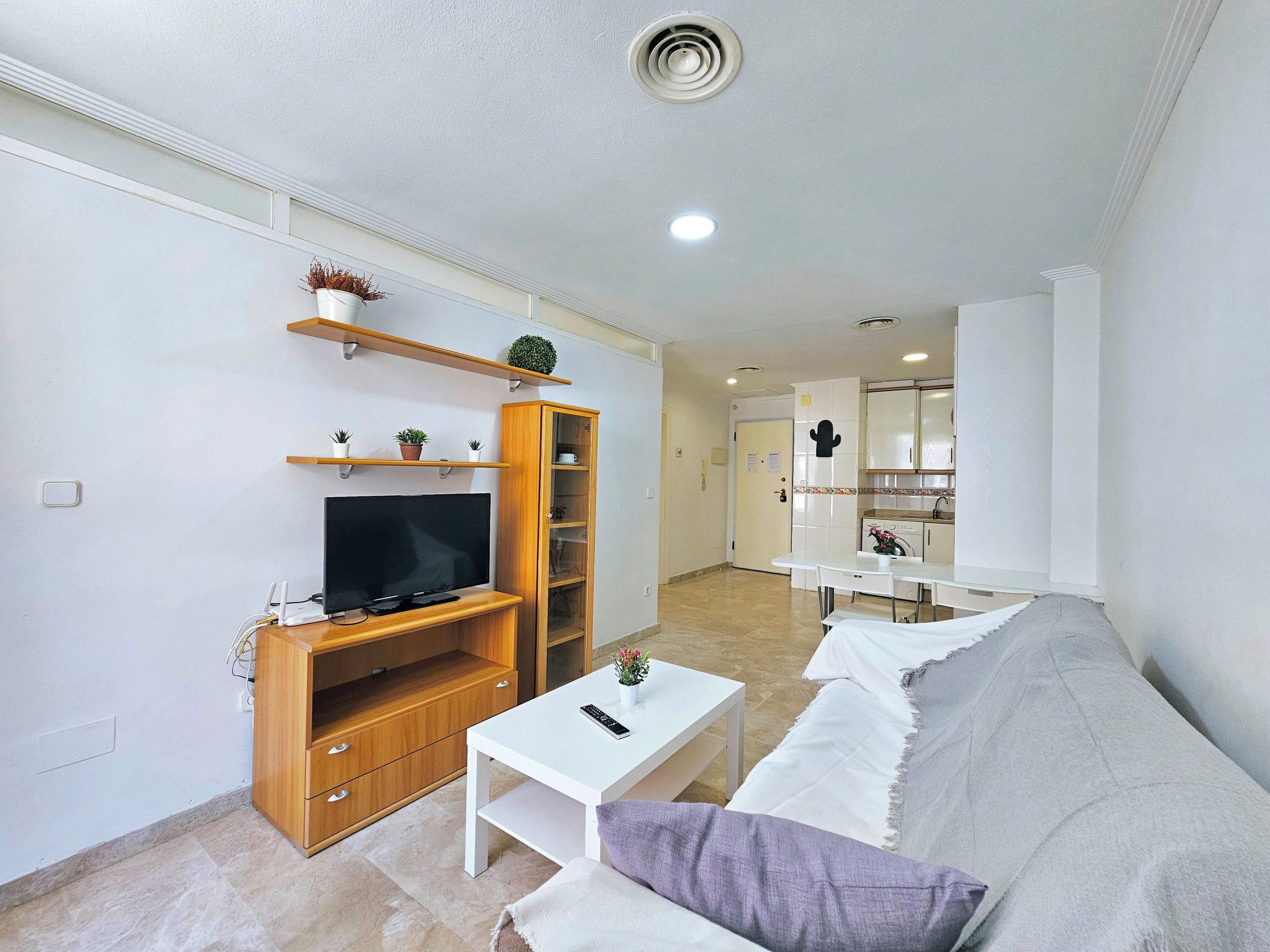 A living room with a TV, wooden entertainment unit, white sofa, and a small table with a remote.
