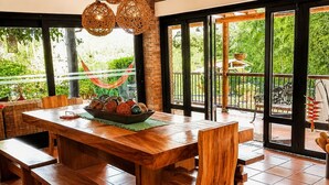 Une table à manger en bois, avec des bancs, entourée de grandes fenêtres et de portes donnant sur un espace extérieur.
