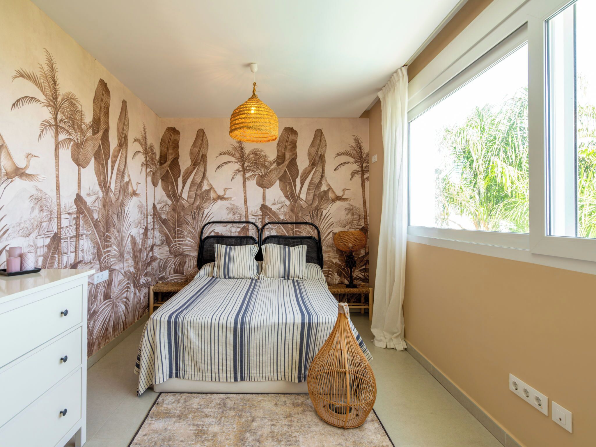 A bedroom with a bed, a dresser, a hanging light fixture, and a large window.