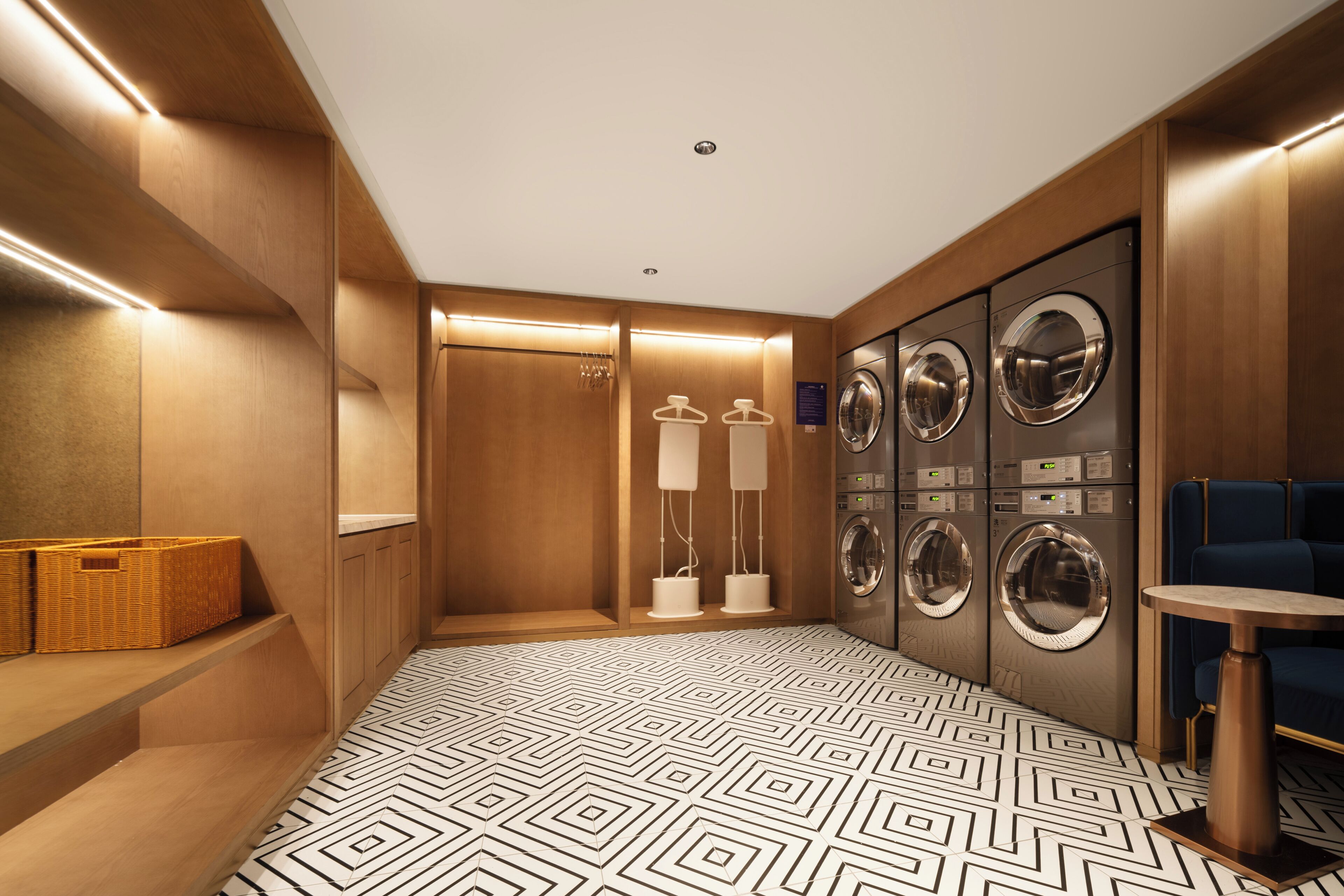A hotel room with a geometric patterned floor, wooden walls, a seating area with baskets, a washing station with dryers, and a small table.
