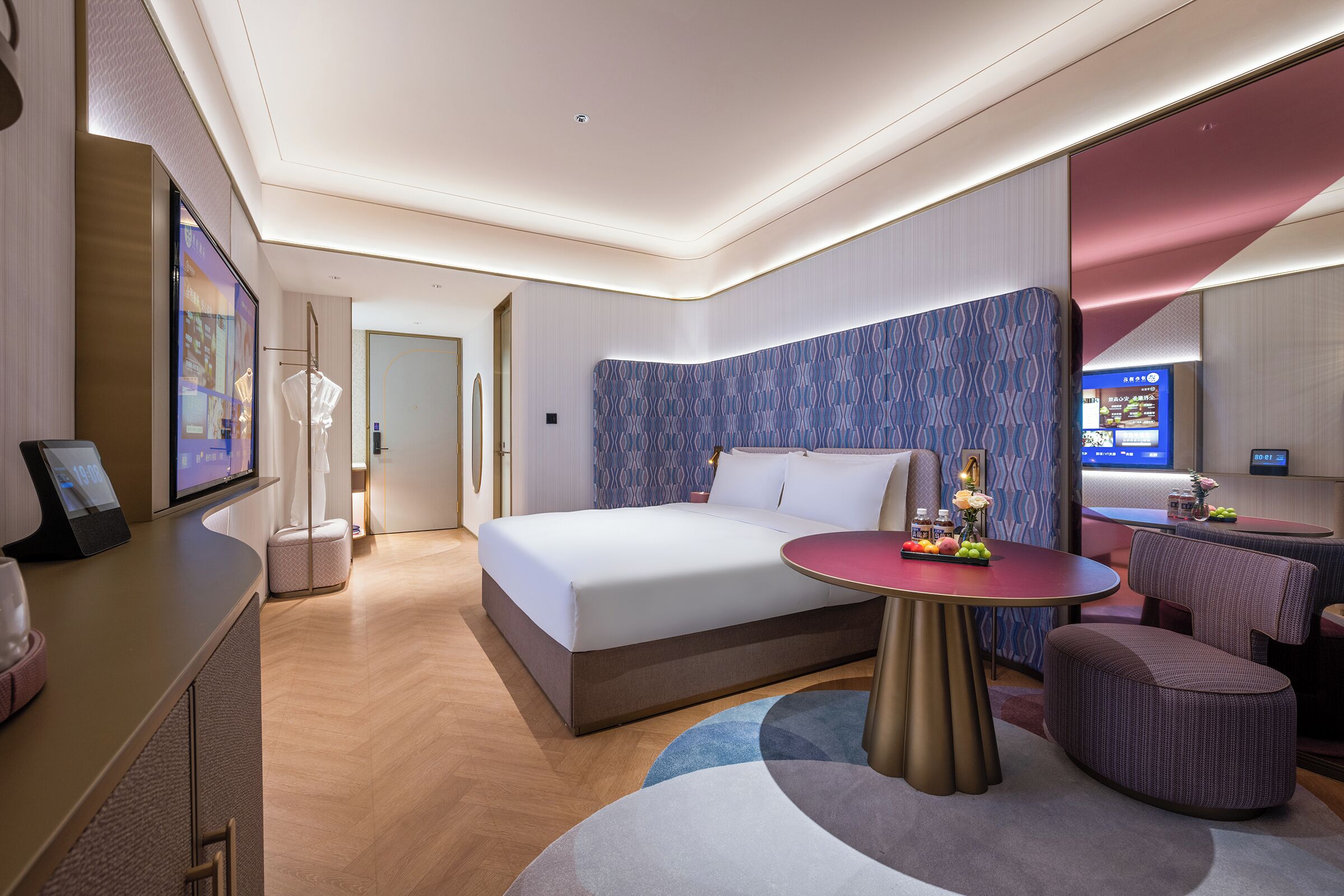 A modern hotel room with a large bed, a round table with fruit, a seating area, and a television.