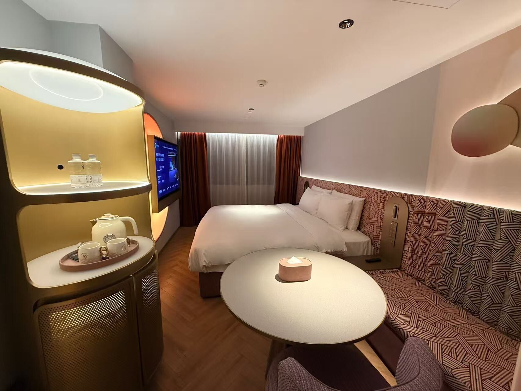 A hotel room with a bed, a sofa, a small table, a TV, and a minibar.