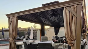 Ein Terrassenbereich im Freien mit Korbmöbeln, einem überdachten Sitzbereich und einem Pool im Hintergrund.