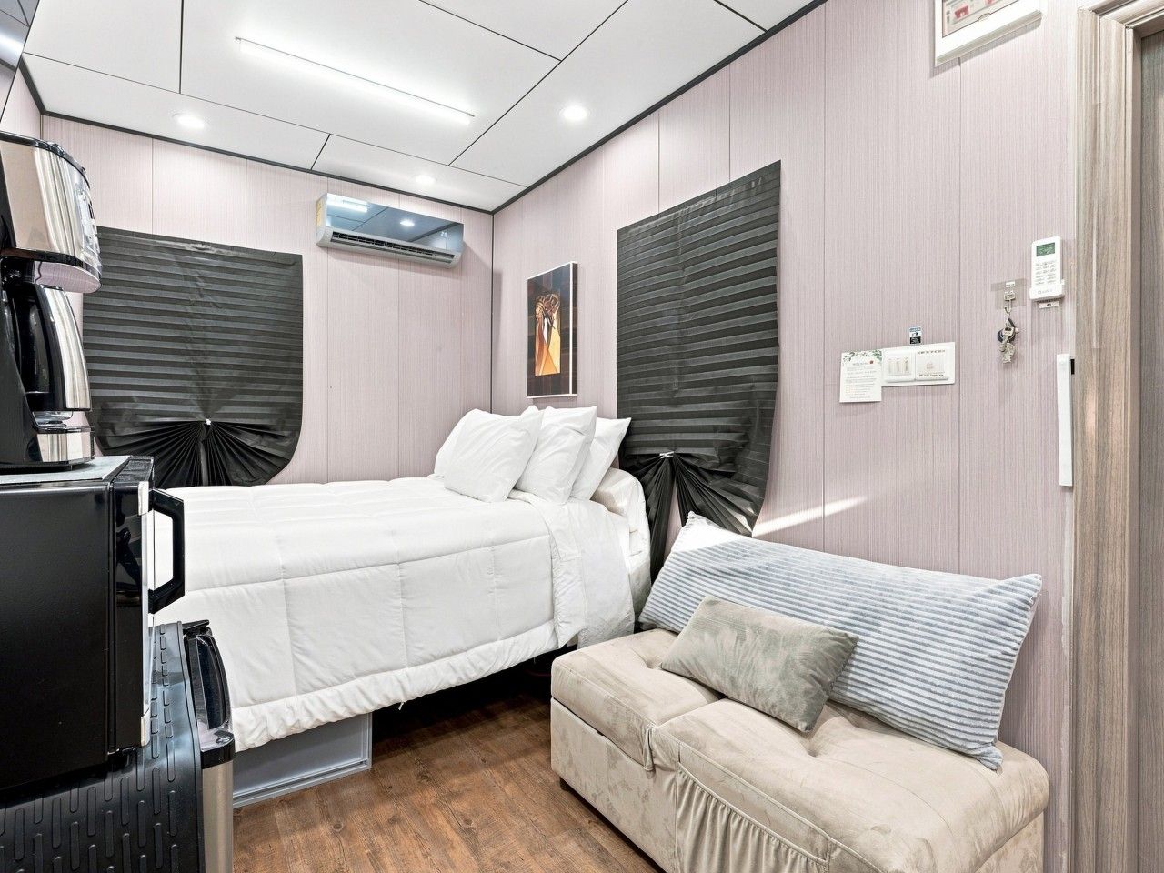 A bedroom with a bed, a sofa, a desk with a computer, and an air conditioning unit.